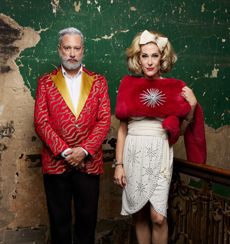 Image of a cis queer gendered man in a red patterned suit with a yellow collar and a them/ they drag performer in a white dress and red fur stole standing in front of a distressed old stucco green wall.