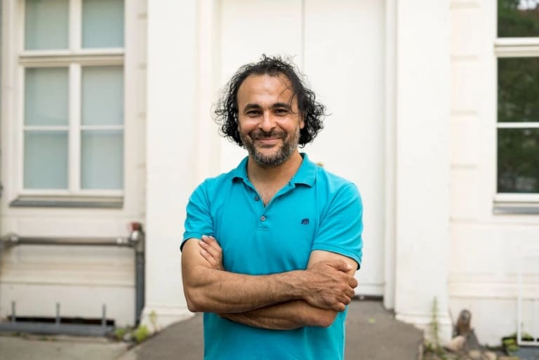 Image of Kader Attia, a man standing in front of a white door with his arms crossed