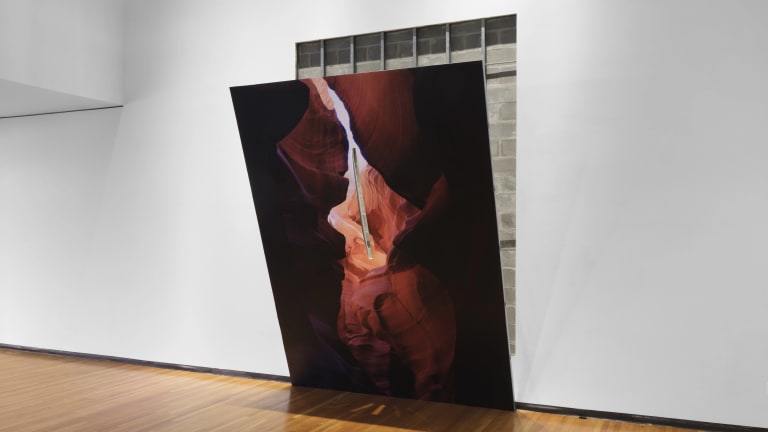 Artwork by Letha Wilson entitled Slit Slot Canyon, 2023. A print of a canyon leans forward off the wall and has a slit in it that looks through to a print of bricks pasted on the wall.Materials are metal studs, drywall, joint compound, digital print on adhesive vinyl, hole in wall. Dimensions are 180" x 110" x 60"