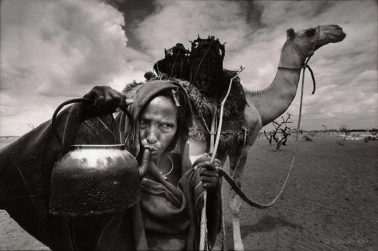 A black and white photo depicting an elderly woman covered in heavy clothes, standing in front of a camel in the desert.