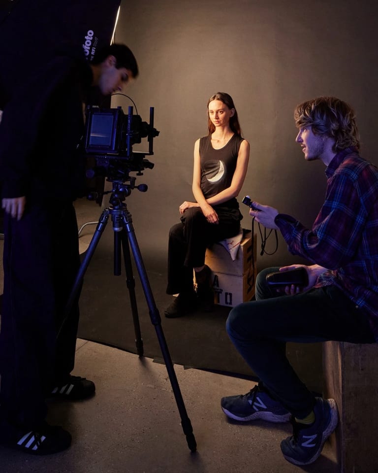 Two students taking a photo of a model in a photography studio with a black back drop.