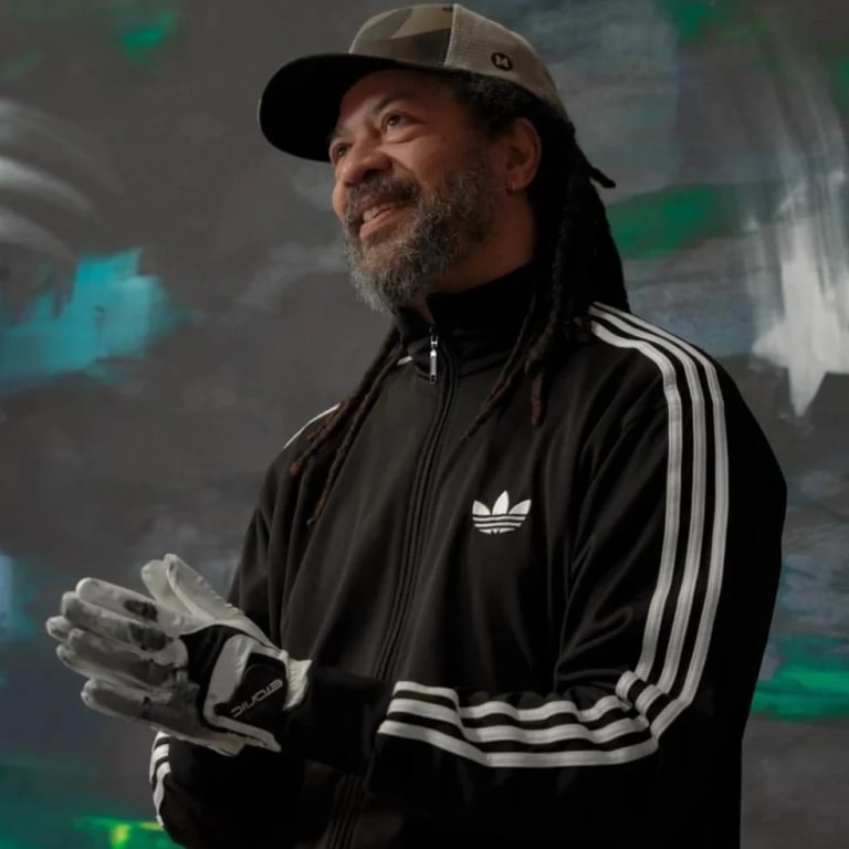 Two images. First: Portrait of a man, Gary Simmons, standing in front of a black wall with white, green and blue paint smudges and starts. The man is wearing a cap, an adidas jacket, and thick gloves. Second: Detail picture of a faded white chalkboard illustration on a dark grey background 