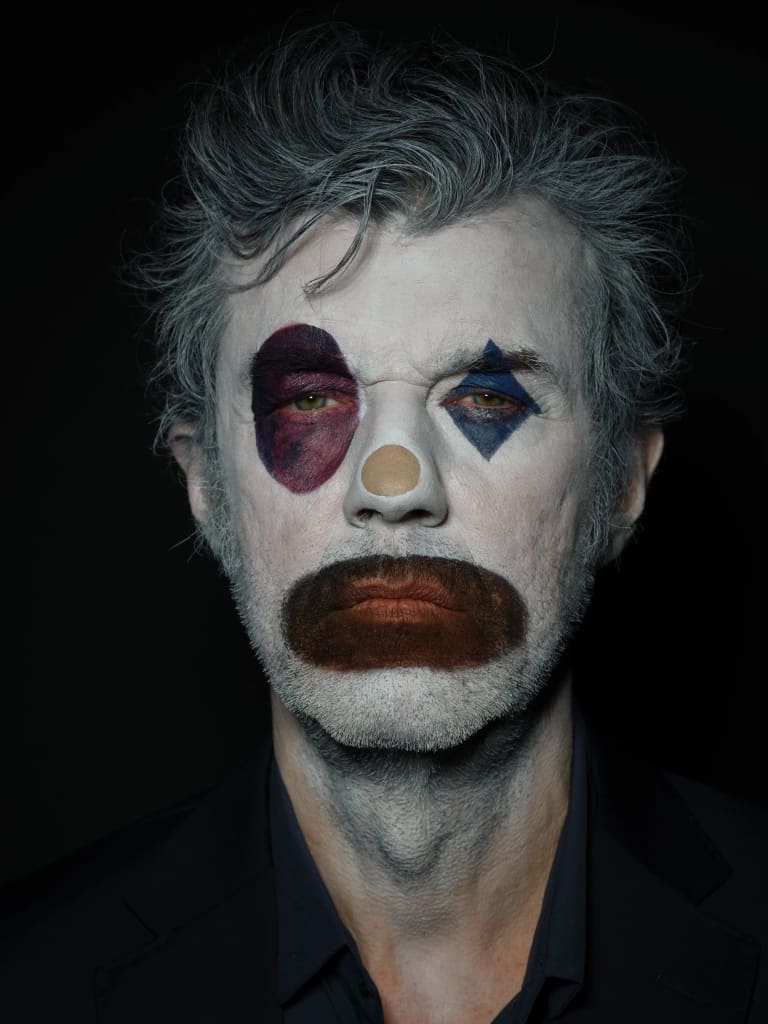 Designer Stefan Sagmeister in clown make-up looks directly at the camera against a dark background.