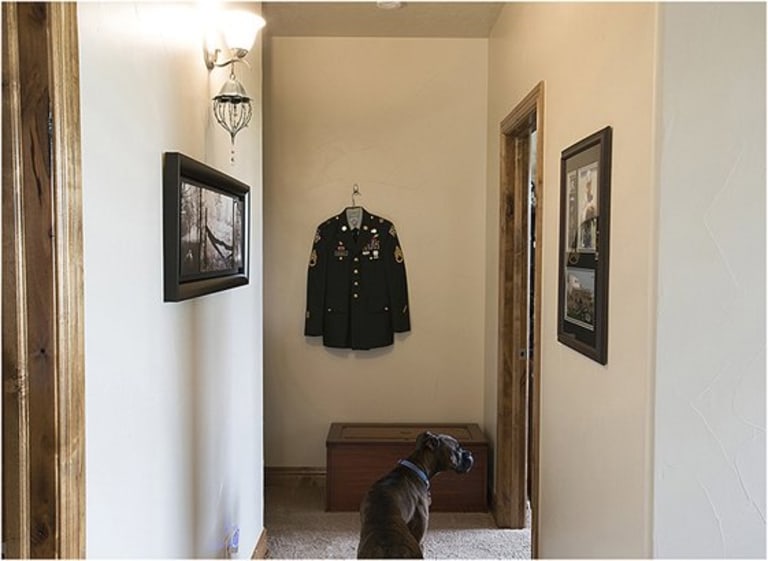 A dog sitting in the hall way in front of an army jacket.
