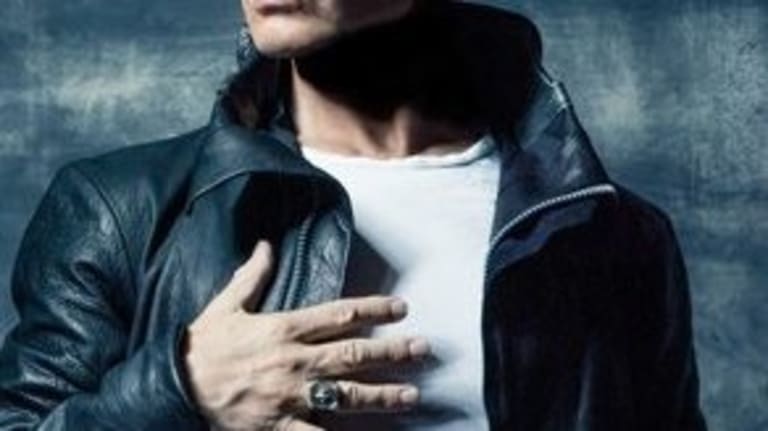 A photograph of a man in a black leather jacket and a plain white t-shirt. He has blond hair and his hand is in front of his chest.
