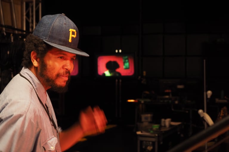 A photo of a man with a beard wearing a dark denim baseball cap with a yellow P on it. It seems as though he is dancing, as his arm is blurred and he is sweaty.