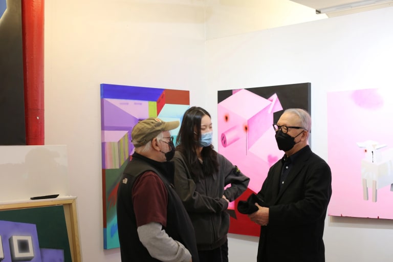 Three people wearing masks stand in a circle in front of multiple paintings 