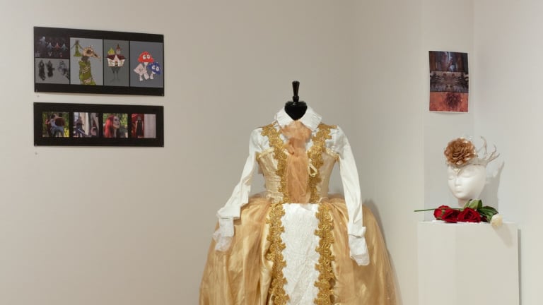 Close-up photograph of ornate white and gold dress on mannequin with prints hung on gallery walls to either side.