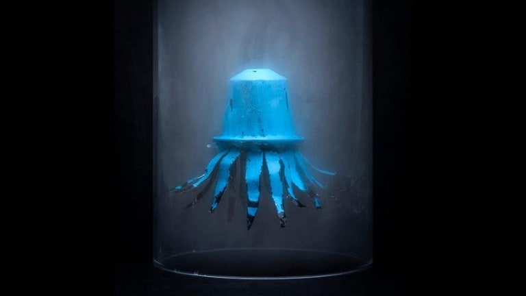 A composite photograph of a clear glass jar filled with fog against a black background. Suspended inside it is a blue coffee capsule with metal tentacles coming from the bottom, making it look like a sea creature.