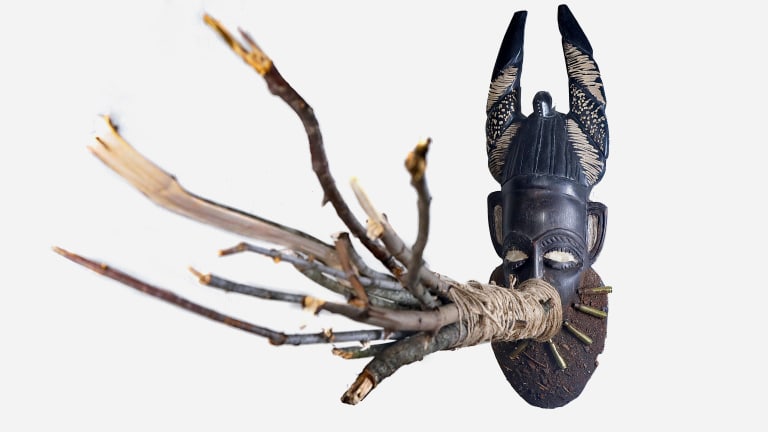 Sculpture using a traditional African mask with a bundle of jagged branches emerging from its mouth.