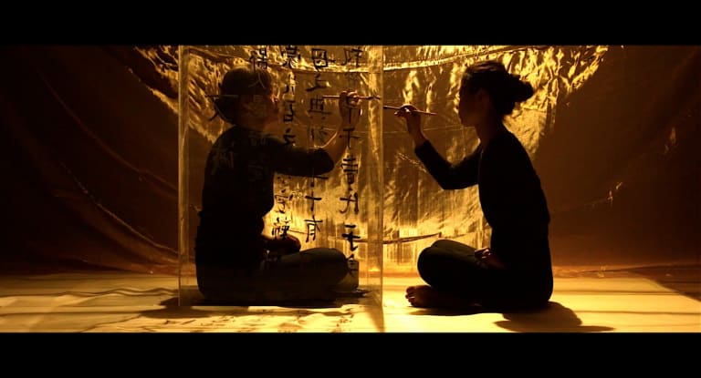 Two women sit cross legged facing each other while they paint Chinese symbols on a glass wall that separates them.