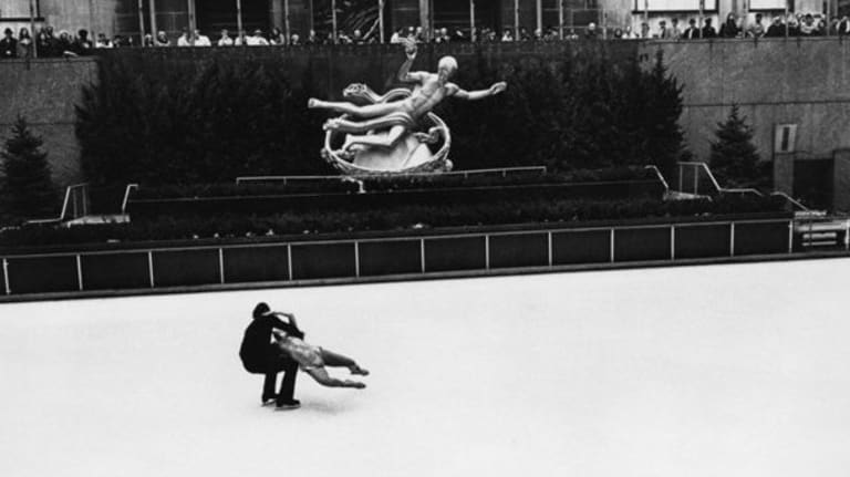 Two people ice-skating on ice with a statue in the background.