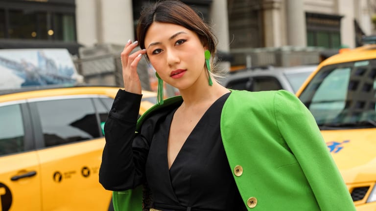 A woman in a black dress and green jacket stands confidently in front of parked taxis on a city street.