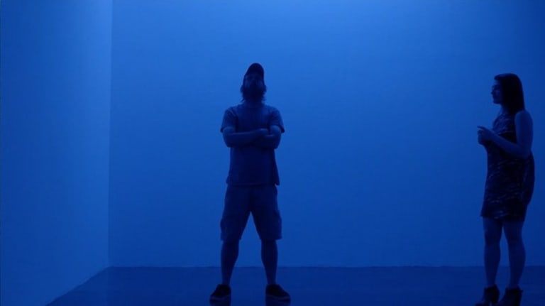 A woman looks at a man in shorts standing in a room illuminated by blue light.