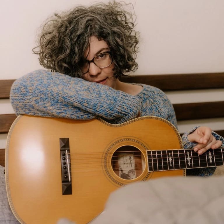 left: an image of a person holding a guitar while sitting in bed; right: an image of a pair of feet wearing pink socks on a bed. 