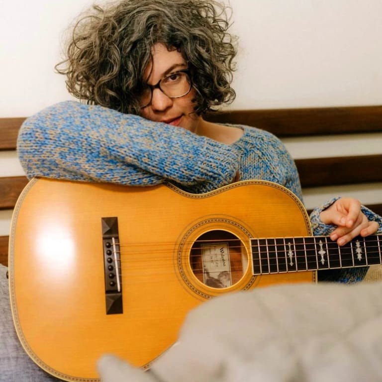 An image of a person wearing in a blue sweater in bed with an acoustic guitar. 