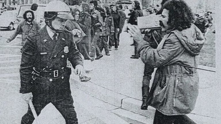 A man with a video camera records a policeman holding a baton as he walks towards him.