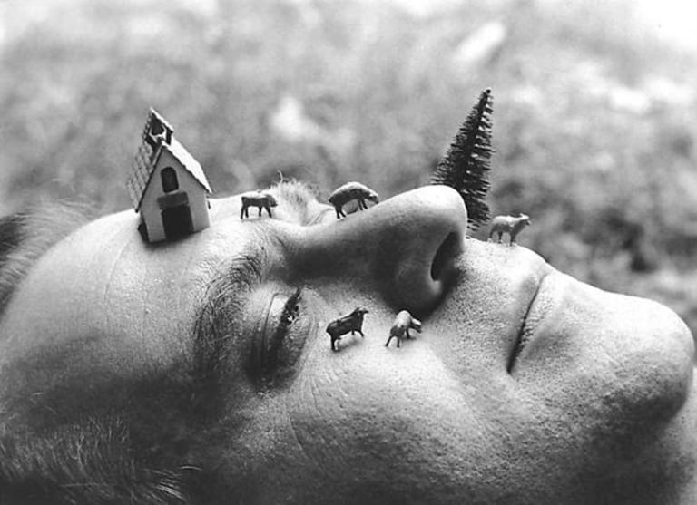Black and white image of a man laying down facing the sky. On his face is a tree and small building surrounded by tiny farm animals.