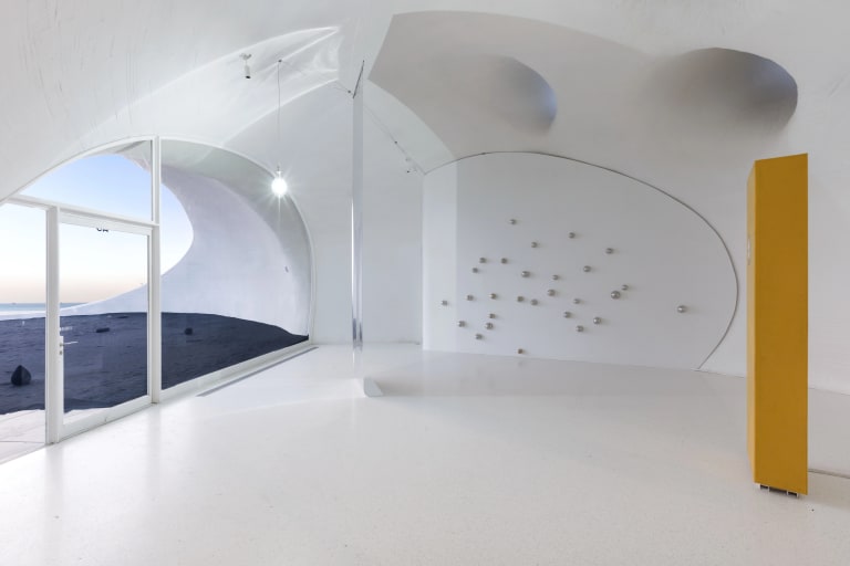 Installation image from the inside of a space with curved architecture, with small chrome orbs mounted on the back wall in a cluster, and a deep yellow rectangular sculpture standing like a column in the right side of the space.