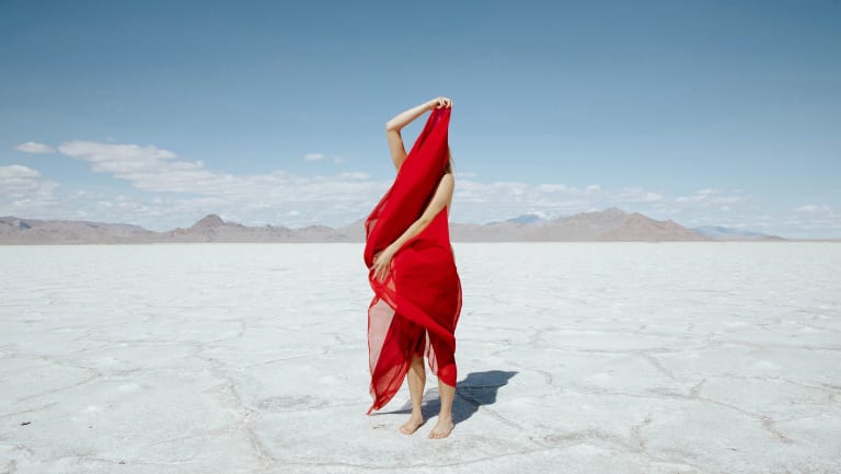 Landscape photograph of a woman holding a red scarf that covers most of her body in the middle of a desert.