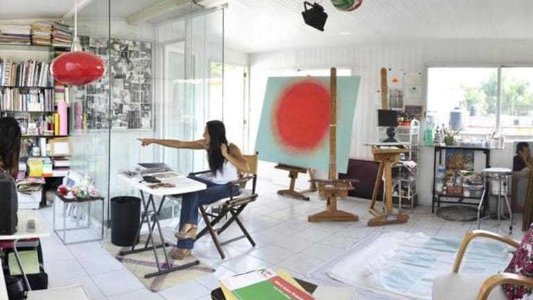 A girl is sitting at a desk and pointing to a stack of books while another girl looks around the corner