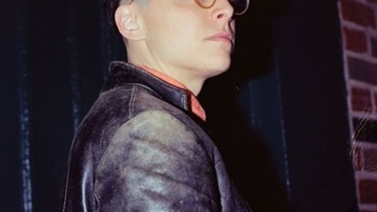 The profile of a dark-haired man wearing a worn leather jacket. The collar of his shirt can be seen and he is also wearing circular glasses.