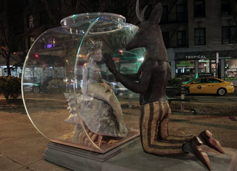 A sculpture on the sidewalk that depicts a mermaid sitting inside a fishbowl on a large hermit crab, looking out the bowl at a large kneeling creature with a mans body and the head of a ram.