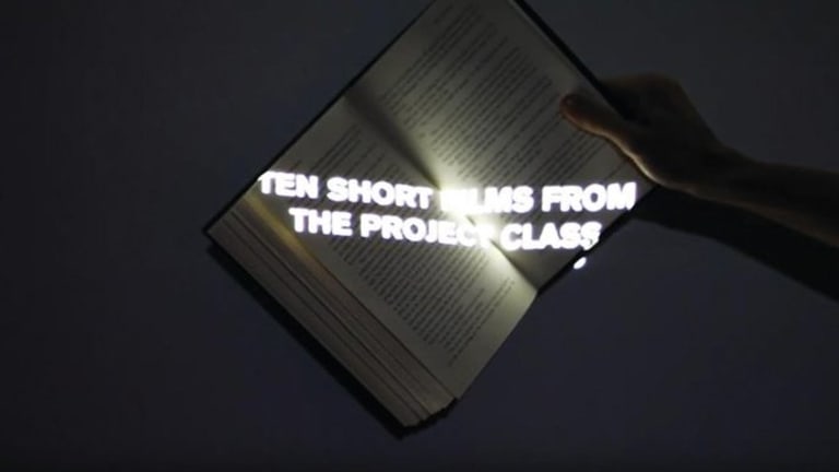 In a darkly lit room and book is held up by a persons hand at a slanted angle. In the center of the book there is a projection of "Ten Short Films From The Project Class" encased in a white light.