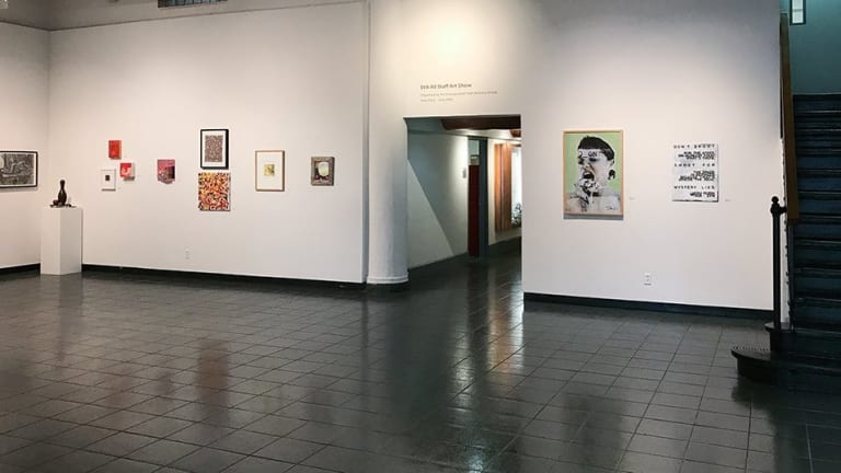 The inside of an art gallery, multiple pieces on display, white background and dark floors