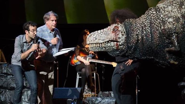 The image is a dinosaur on stage with people. Two men have microphones and one person has a guitar.