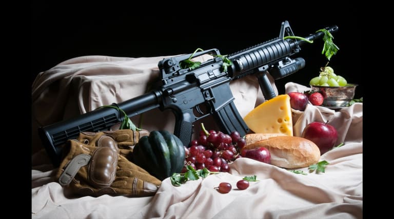 Machine gun displayed in a surreal still art scene with fruit, cheese, bread, and gloves