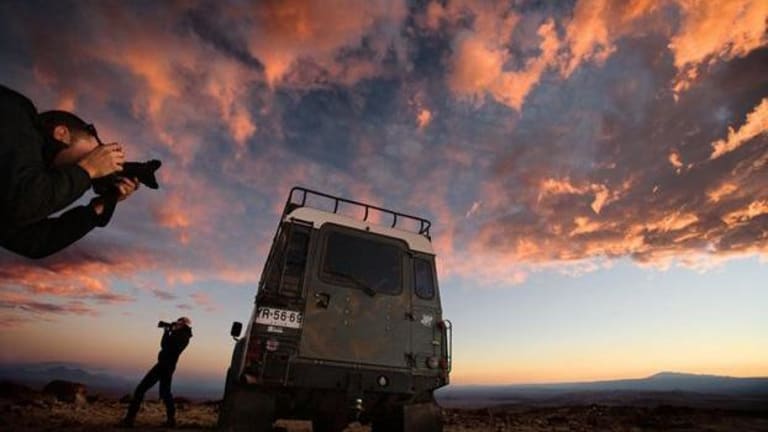 Two photographers are in the desert. There is a jeep, and the sky is pink and blue.