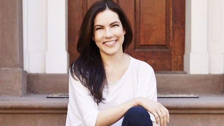 A photo of a smiling woman with dark long hair, a white t-shirt and jeans sitting on some steps