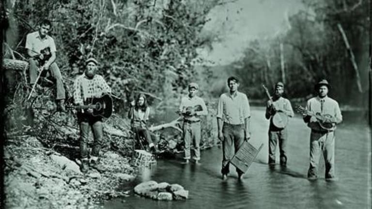 Seven men with musical instruments standing in a creek.