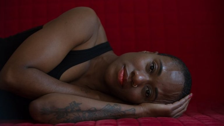 There is a black woman with short cropped hair laying on her side on a red velvet sofa. She is wearing a black tank top and nose ring. She has a tattoo on her left arm