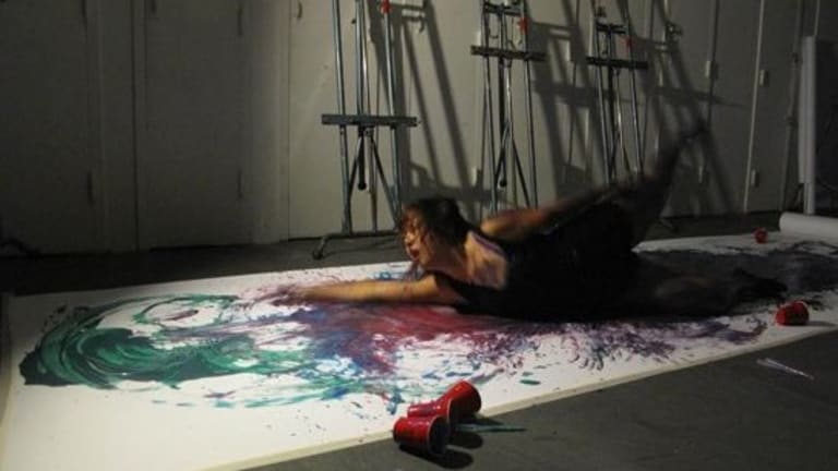 A room with a woman wearing all black, laying on her side on the floor. Under her is a white canvas with paint all over.