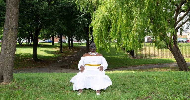 A woman in a white dress wearing a black mask sits in the middle of a green park space.