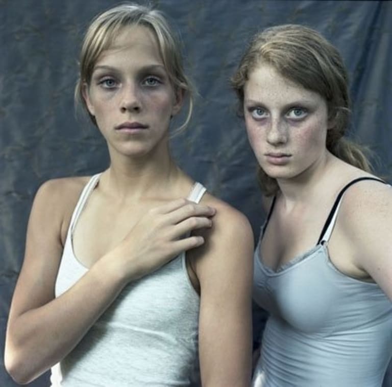 Two women in tank tops stand in front of a blue backdrop.