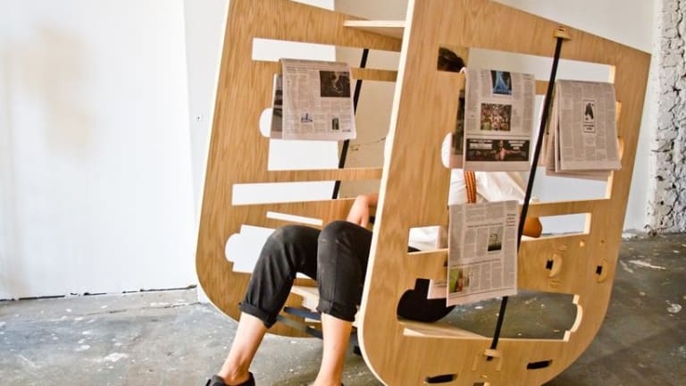 Person sitting in abstract rocking chair with newspapers hanging off of side of chair.