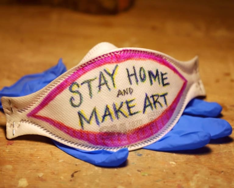 A face mask sits atop some rubber gloves. Written on the face mask is the phrase "Stay home and make art"