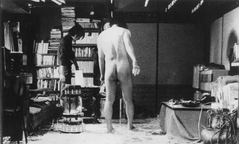 black and white photo of two men, one clothed one naked, standing in a room lined with books