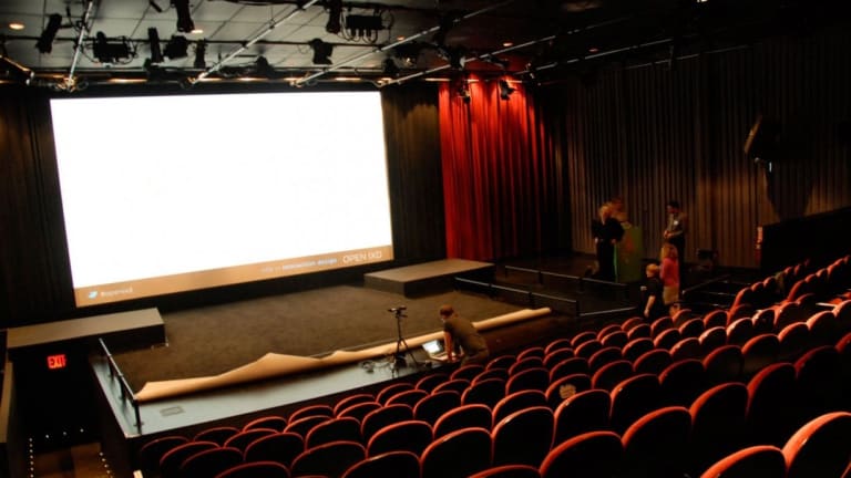 A white screen shines brightly in an auditorium with a few technicians and empty red seats.