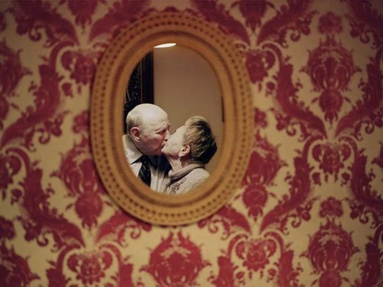 An older man and woman kissing in an oval mirror on a wallpapered wall.