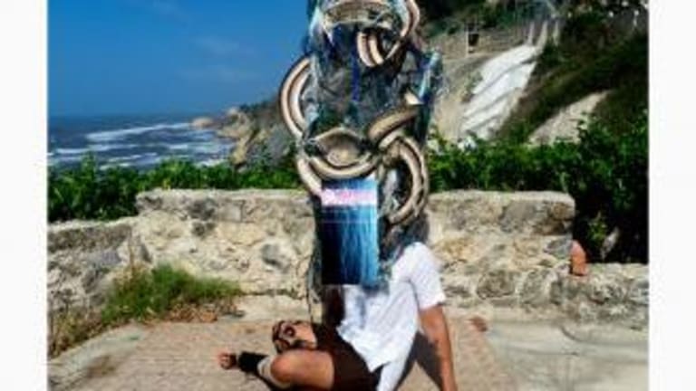 A man is seated cross legged on a straw mat, behind a sculpture of black and white ribbons with blue feathers.