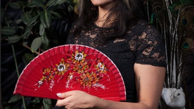There is a woman sitting in a black dress with lace sleeves. She is holding a red fan with a floral design in front of her chest.
