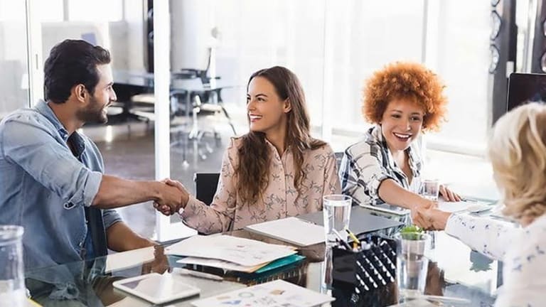 Photo of four people sitting at a conference table, each shaking hands with one of the others. There are various office supply items neatly on the table in front of the people.