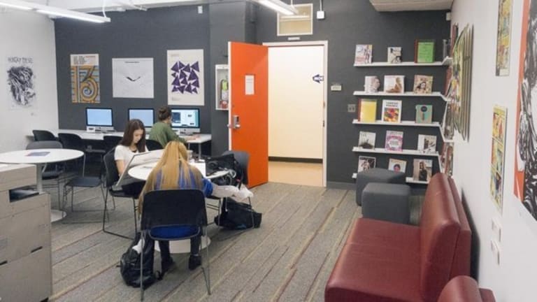 A break room with various posters covering the walls. In the center of the room are two women sitting on laptops, with a third man farther away from them on a computer close to a wall.