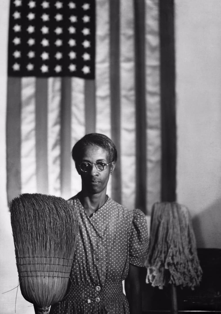 A Black woman stands in front of the American flag holding a broom and a mop.
