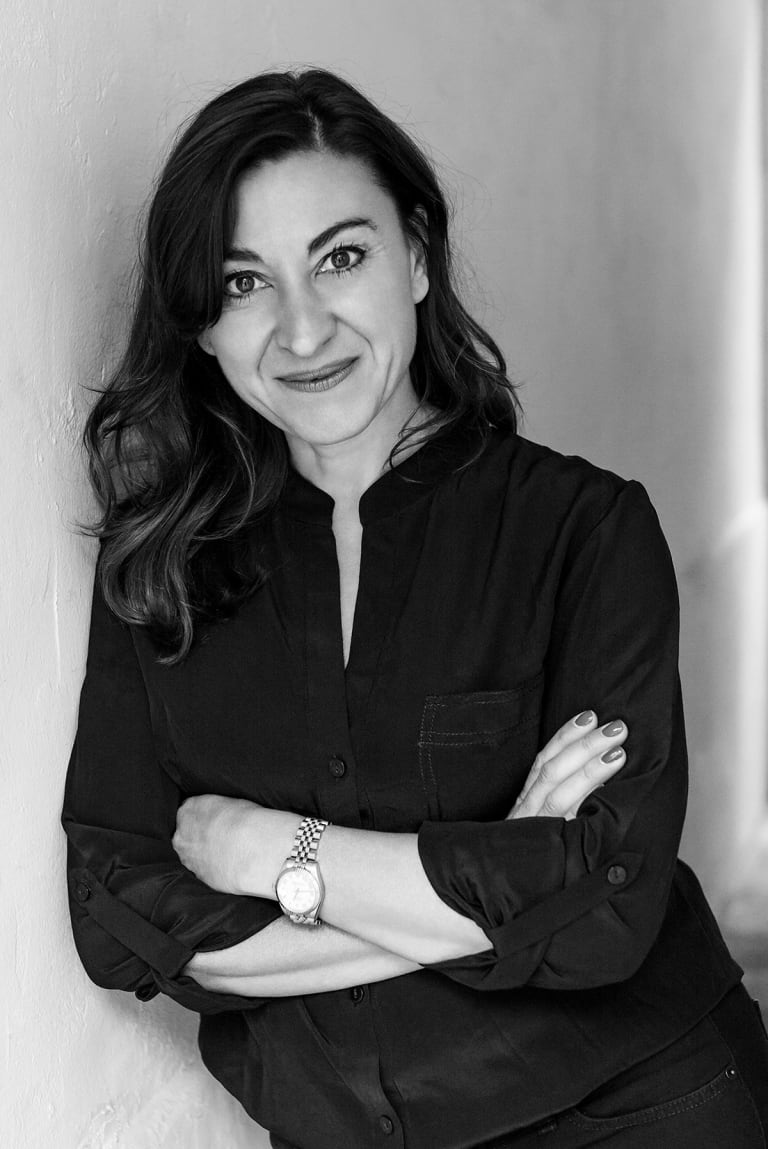 Black and white portrait of Lynsey Addario learning against a wall with arms crossed, smiling.