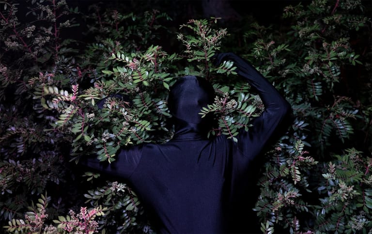 A man in a black bodysuit stands in front of a tree and stretches his arms back to hug the tree behind him.